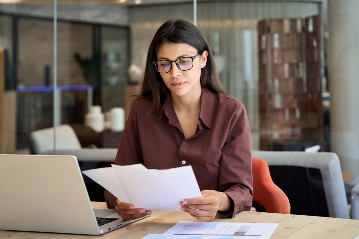Focused businesswoman working on a laptop and reviewing a financial document in an office, organizing records for a self-employed loan application