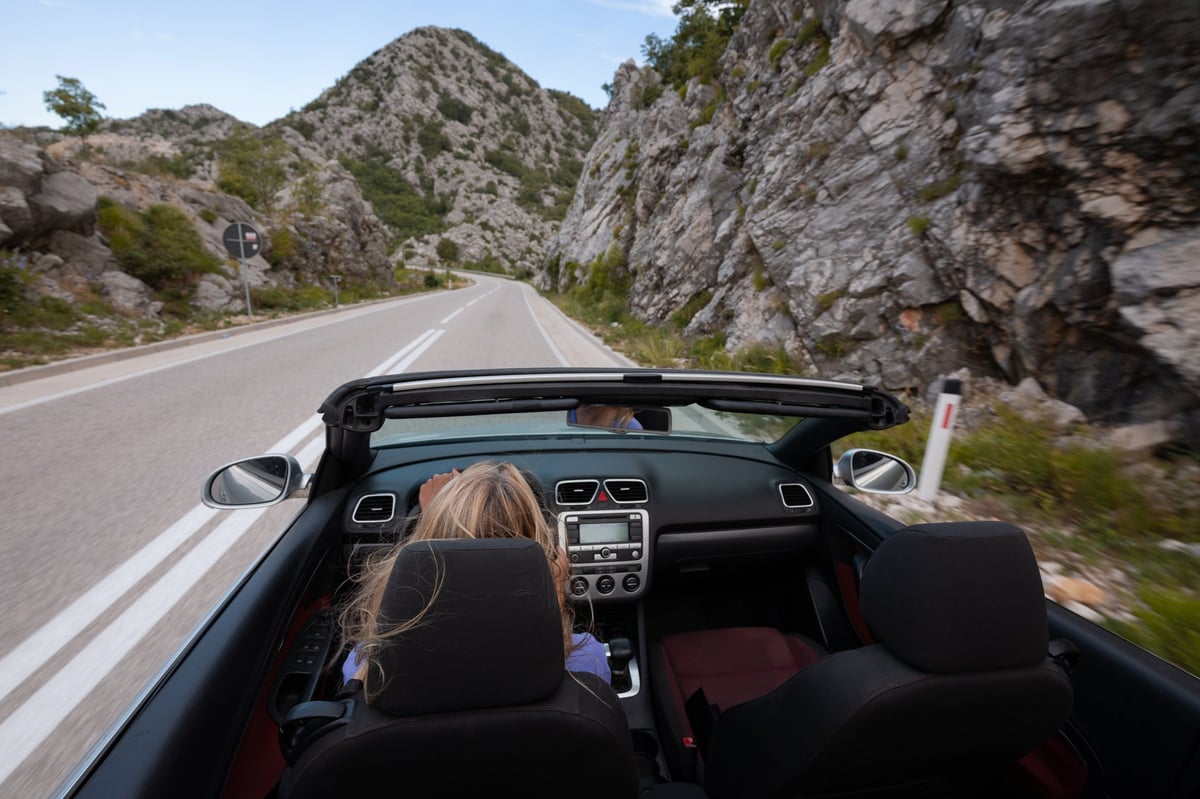 Young woman driving convertible along scenic ocean coast and mountain road at sunset, carefree summer vacation, travel freedom and adventure on coastal highway