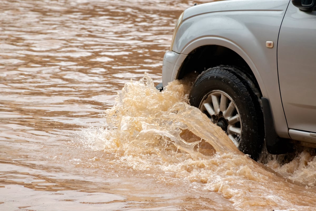 Dark yellow floodwater covers a rural road, hindering commuters, illustrating flood damage risks for used car buyers