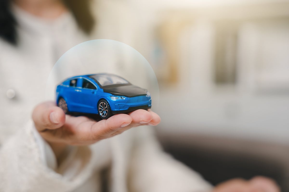 Woman hand holding blue toy car under protective shield for auto insurance service, protecting vehicle from accident or damage with secure commercial transport contract.