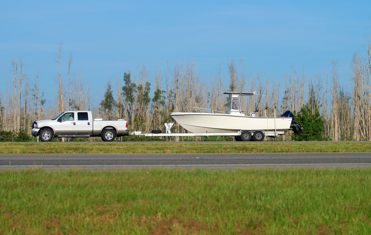 Truck towing a large boat on a trailer along a highway, with bare trees and clear blue sky in the background.