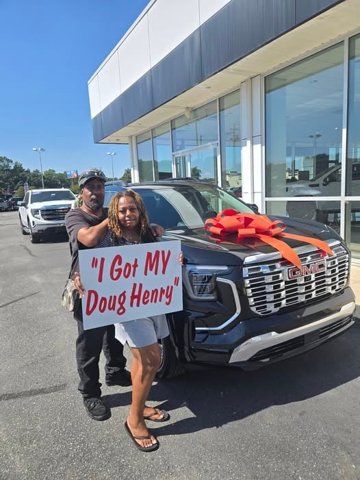 Two people holding a sign that says I Got MY Doug Henry stand next to a new dark-colored GMC truck with a red bow at a dealership.