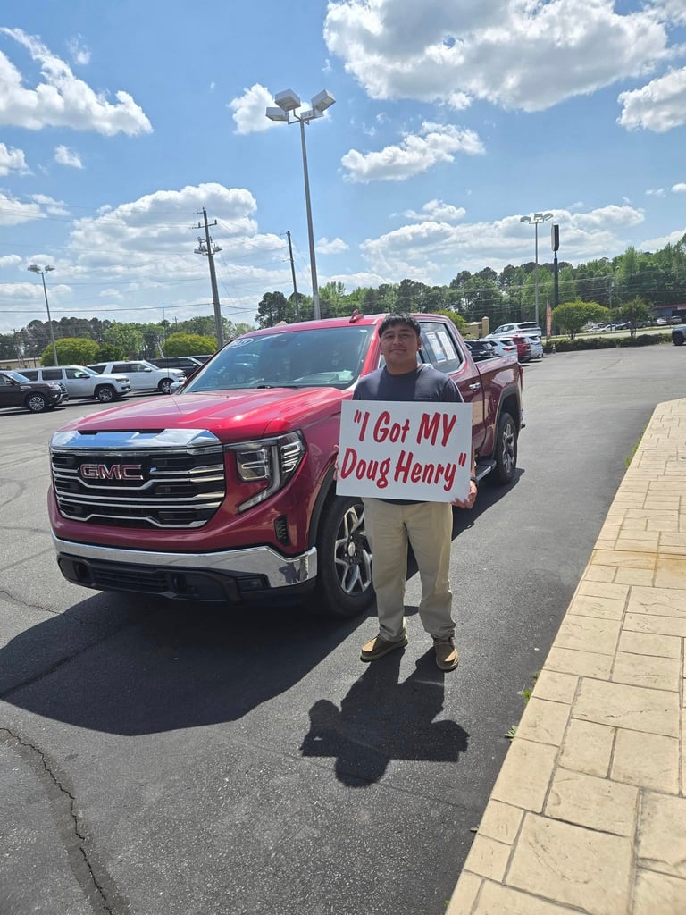 Man holding a sign reading I Got MY Doug Henry stands in front of a red GMC truck in a parking lot on a sunny day