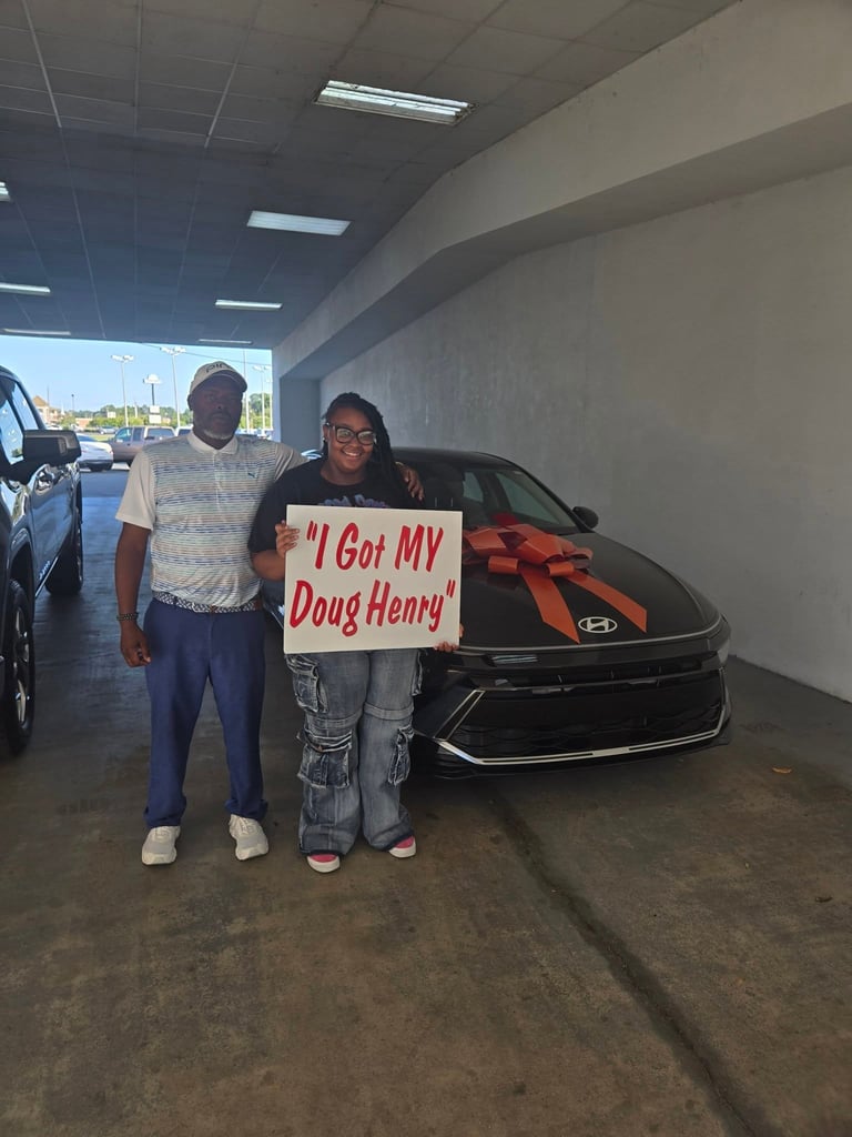 Two people stand next to a black Hyundai vehicle in a parking garage, holding a sign that reads I Got MY Doug Henry