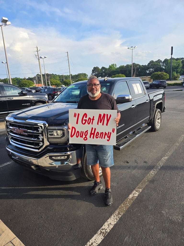 Man holding a sign that says I Got MY Doug Henry standing in front of a black pickup truck at a dealership parking lot