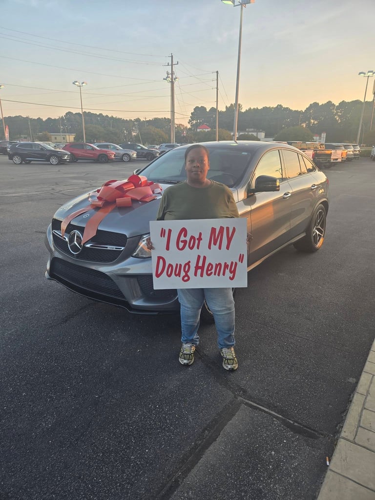 Person holding I Got MY Doug Henry sign standing in front of a new Mercedes-Benz with a red bow in a parking lot