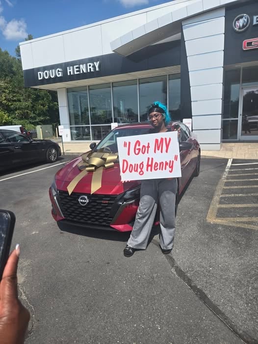 Man holding I Got MY Doug Henry sign in front of maroon vehicle at Doug Henry dealership