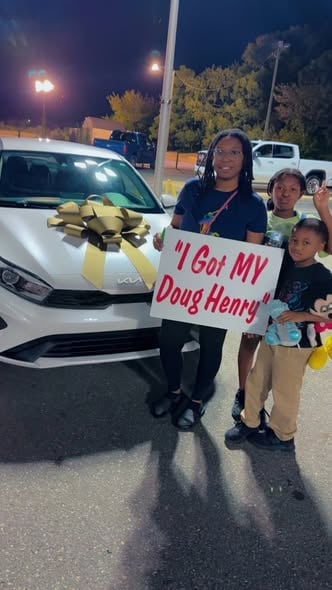 Woman and children pose with a white car decorated with a gold bow at night, holding a sign that reads I Got My Doug Henry