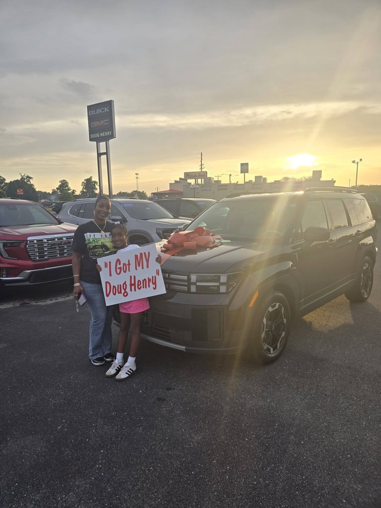 Person holding I Got MY Doug Henry sign stands in front of new SUV at car dealership lot during sunset with city skyline in background
