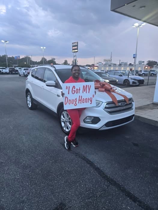 Person in red outfit holds sign reading I Got MY Doug Henry while standing next to white SUV in dealership parking lot