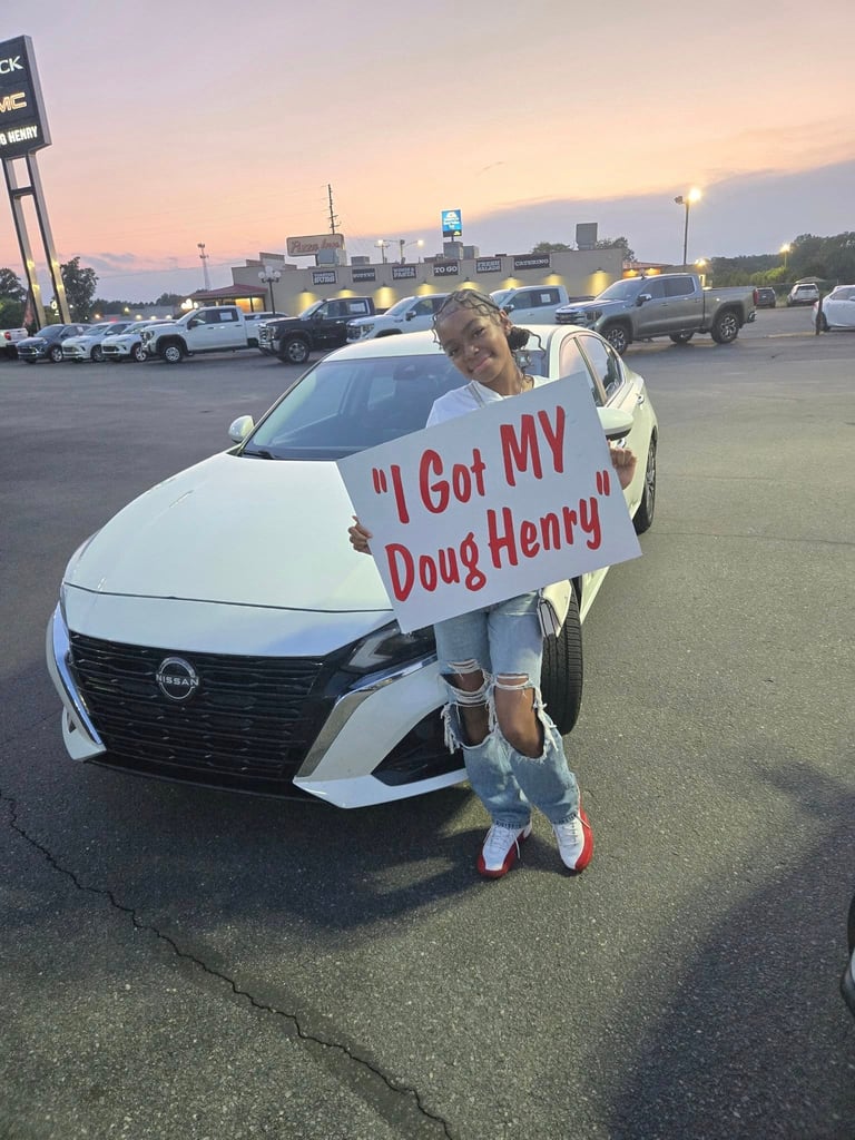 Customer holding sign reading I Got MY Doug Henry standing next to white Nissan sedan at car dealership lot at dusk