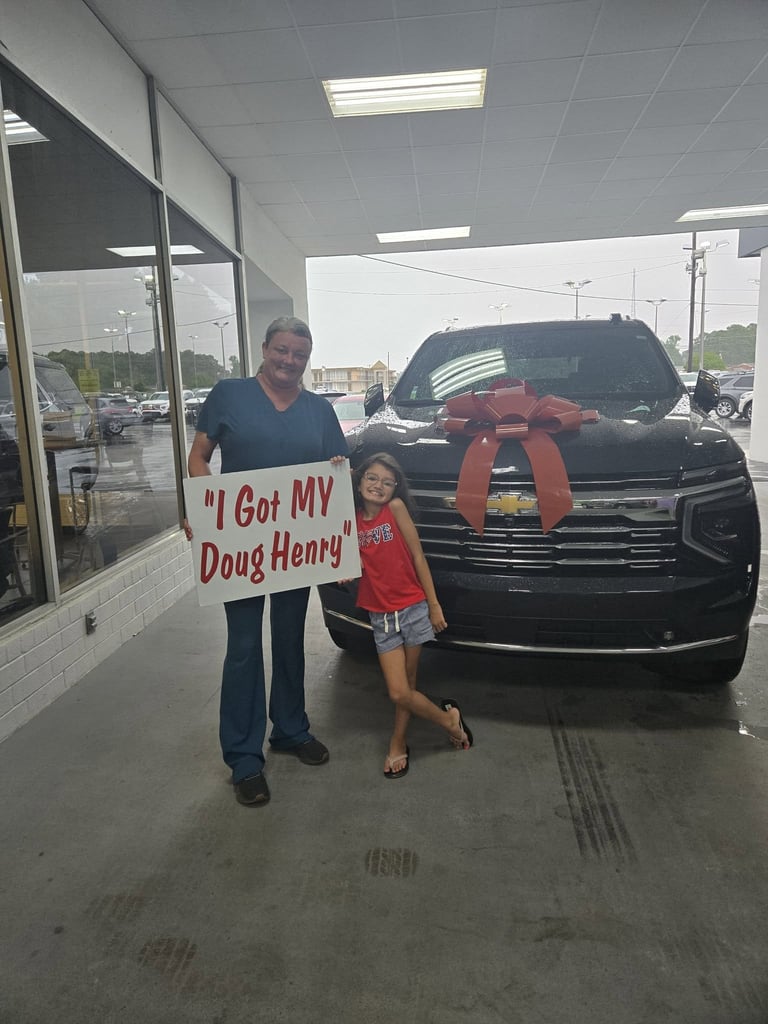 Adult and child hold sign I Got MY Doug Henry in front of new black truck with red bow at dealership