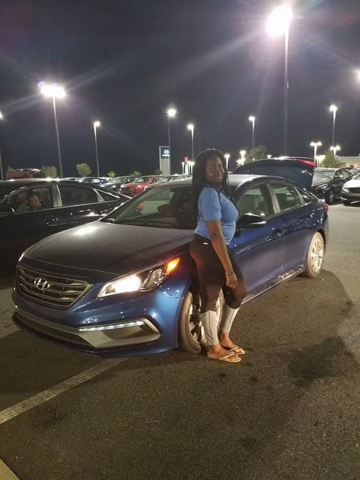 Woman posing next to a blue Hyundai sedan in a nighttime parking lot illuminated by street lights