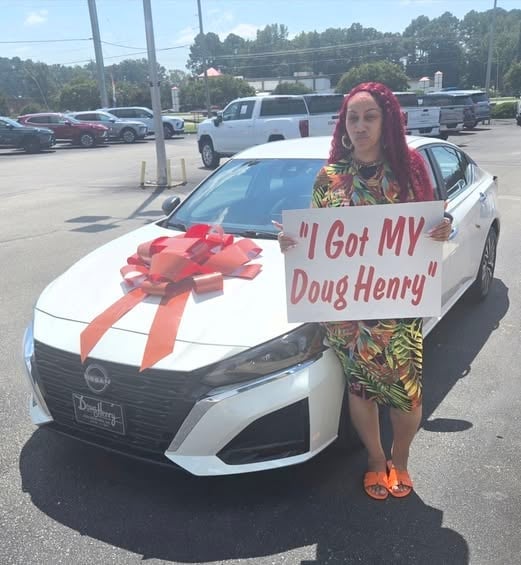 Woman with red hair standing next to a white Nissan with a red bow and sign reading I Got MY Doug Henry
