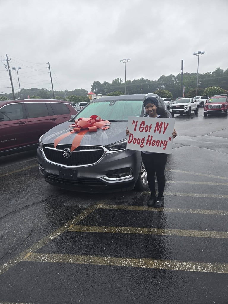 Person holding a sign that reads I Got MY Doug Henry stands in front of a silver SUV with a red bow in a car dealership parking lot