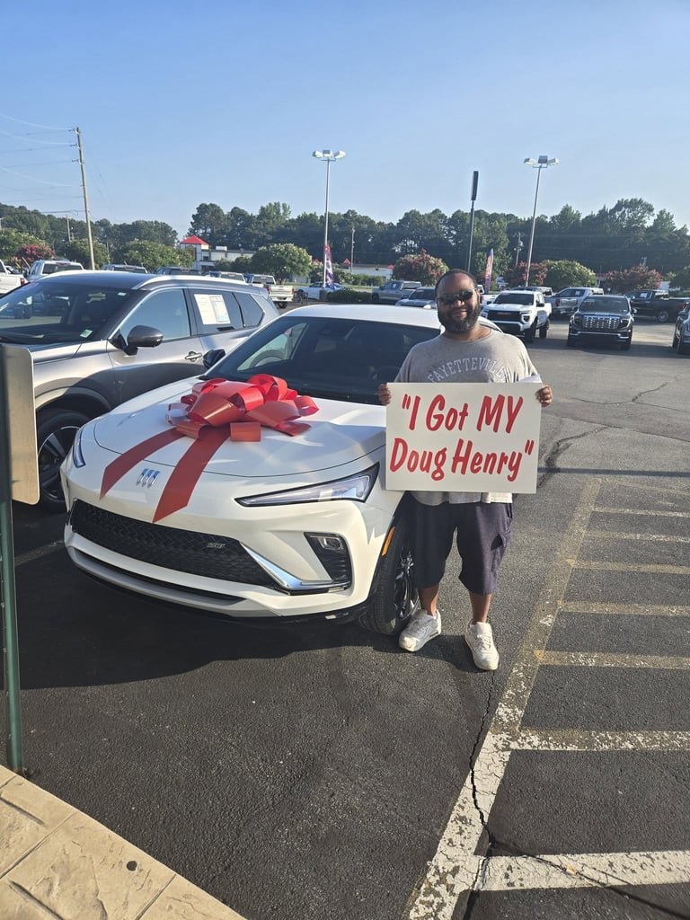Man holding sign reading I Got MY Doug Henry next to a white Chevrolet Corvette with a red bow in a dealership lot