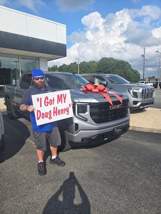 Man holding I Got My Doug Henry sign at car dealership lot with new trucks and red bow