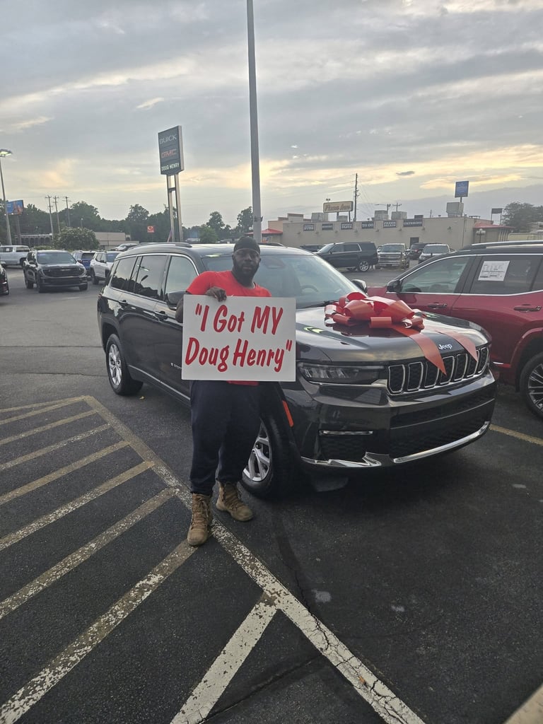 Person holding sign in parking lot stands in front of black SUV with red bow, text reads I Got MY Doug Henry
