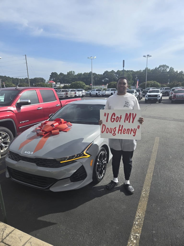 Person holding an I Got My DougHenry sign standing next to a silver sports car with a red bow in a parking lot