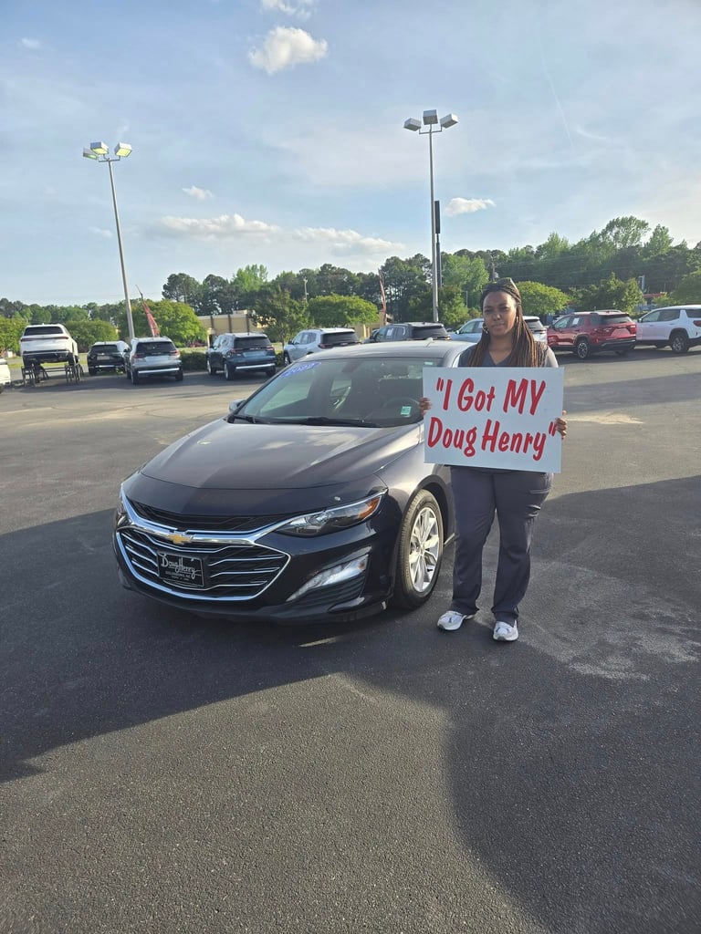 Person holding a sign reading I Got MY Doug Henry stands in front of a black Chevrolet sedan in a parking lot