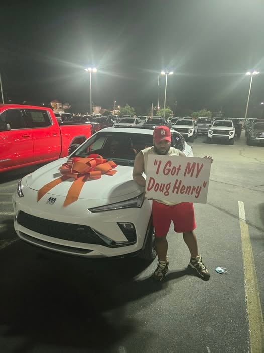 Man standing next to white Camaro with large red bow in parking lot at night, holding sign reading I Got MY Doug Henry