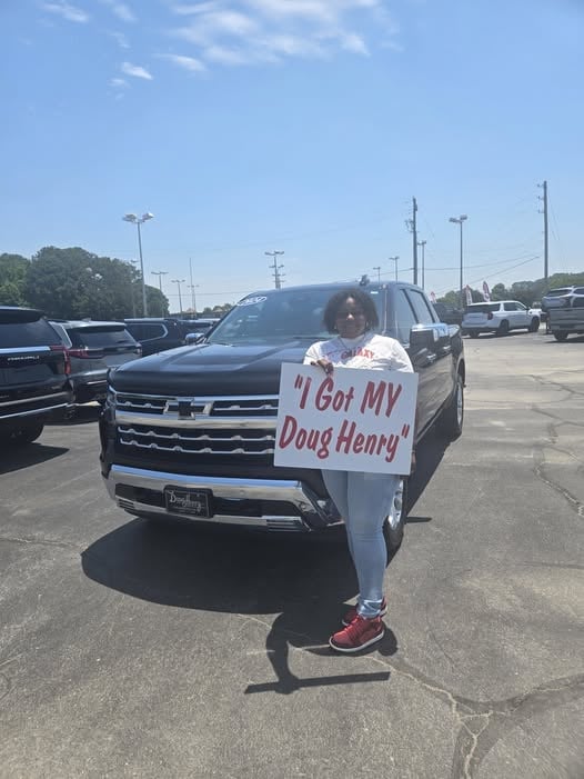 Person standing in front of a dark Chevrolet SUV in a parking lot holding a sign that says I Got MY Doug Henry