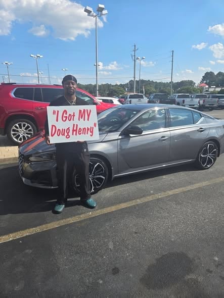 Man holding a sign saying I Got MY Doug Henry standing in front of a silver sedan at a car dealership lot
