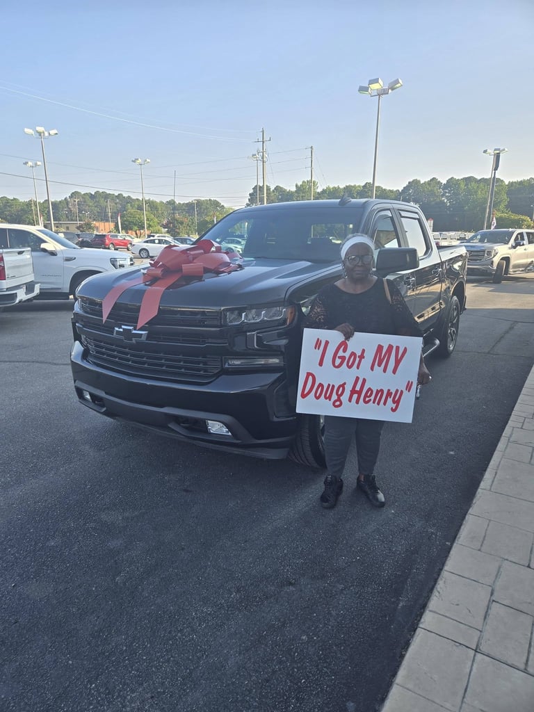 Person holding a sign reading I Got MY Doug Henry stands in front of a black pickup truck with a large red bow in a dealership parking lot.
