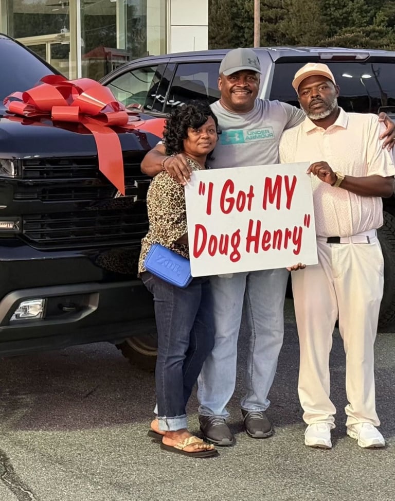 Three people stand in front of a black car with a red bow, holding a sign that says I Got MY Doug Henry