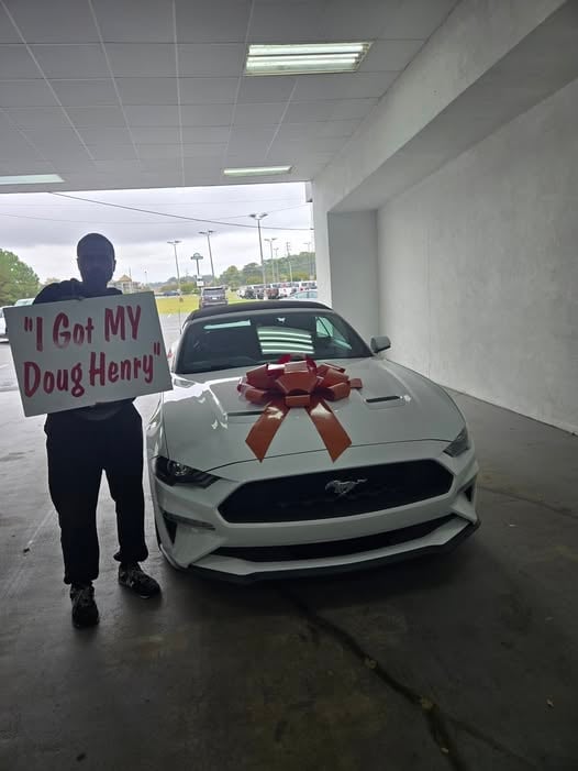 Person holding sign reading I Got MY Doug Henry stands next to white Ford Mustang with large red bow in covered garage