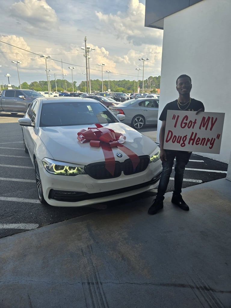 Man holding sign that reads I Got MY Doug Henry stands next to white BMW car with red bow in parking lot