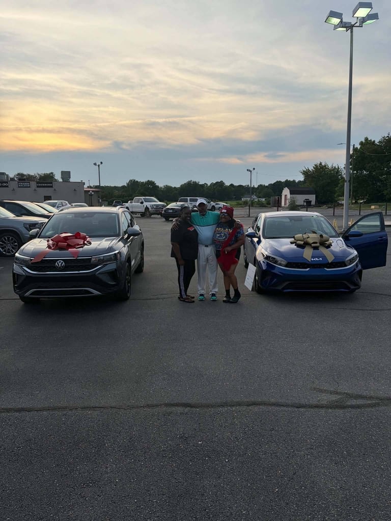 Three people posing between two cars in a parking lot at dusk with tall light poles and buildings visible in the background