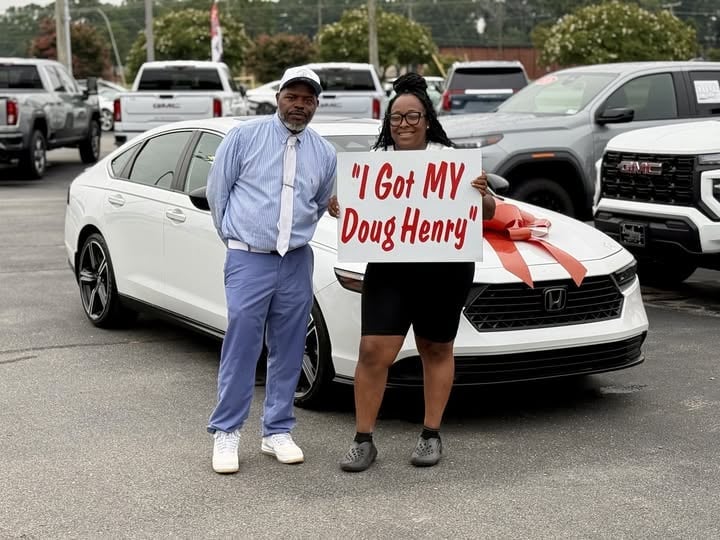 Two people holding a sign reading I Got MY Doug Henry stand in front of a white Honda sedan in a parking lot
