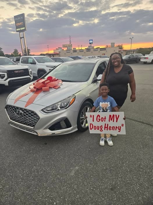 Woman and child posing with new white car with red bow at dealership at sunset, child holds sign saying I Got MY Doug Henry