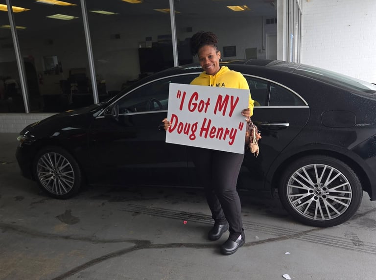 Person in yellow shirt holding I Got MY Doug Henry sign next to a black luxury sedan in a showroom