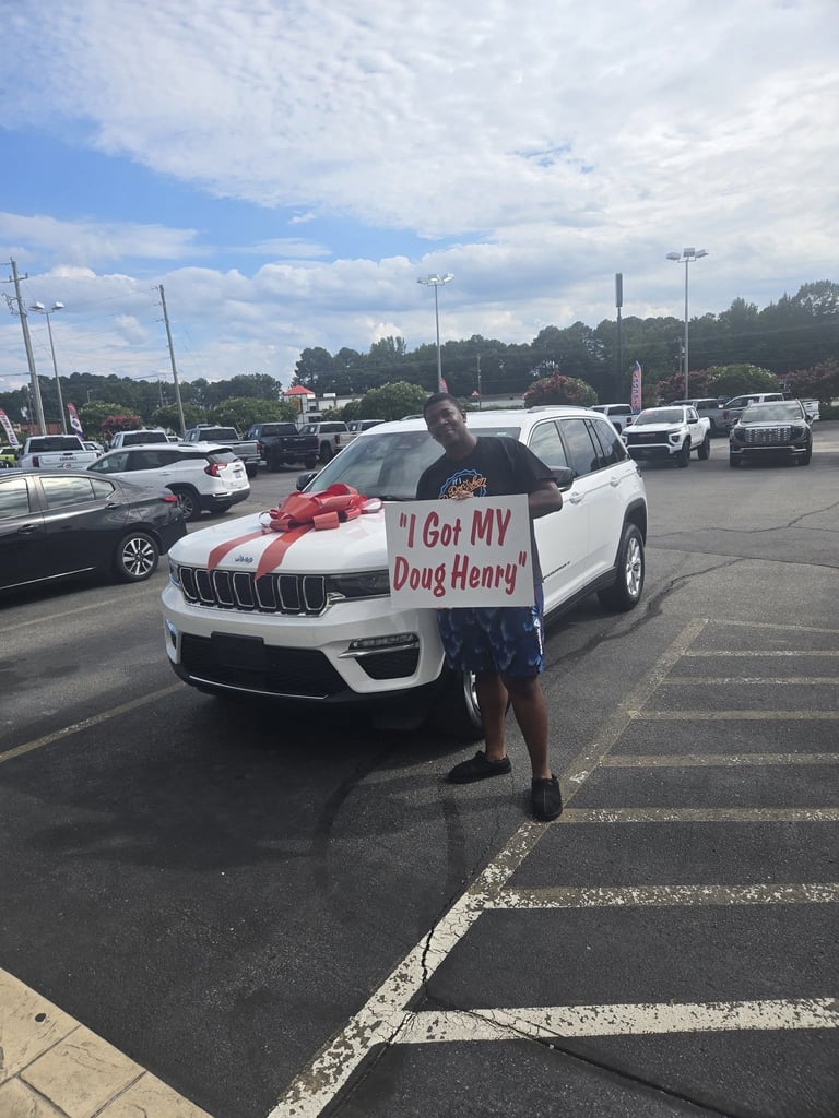 Man posing with white Jeep SUV decorated with red bow and I Got MY Doug Henry sign in parking lot