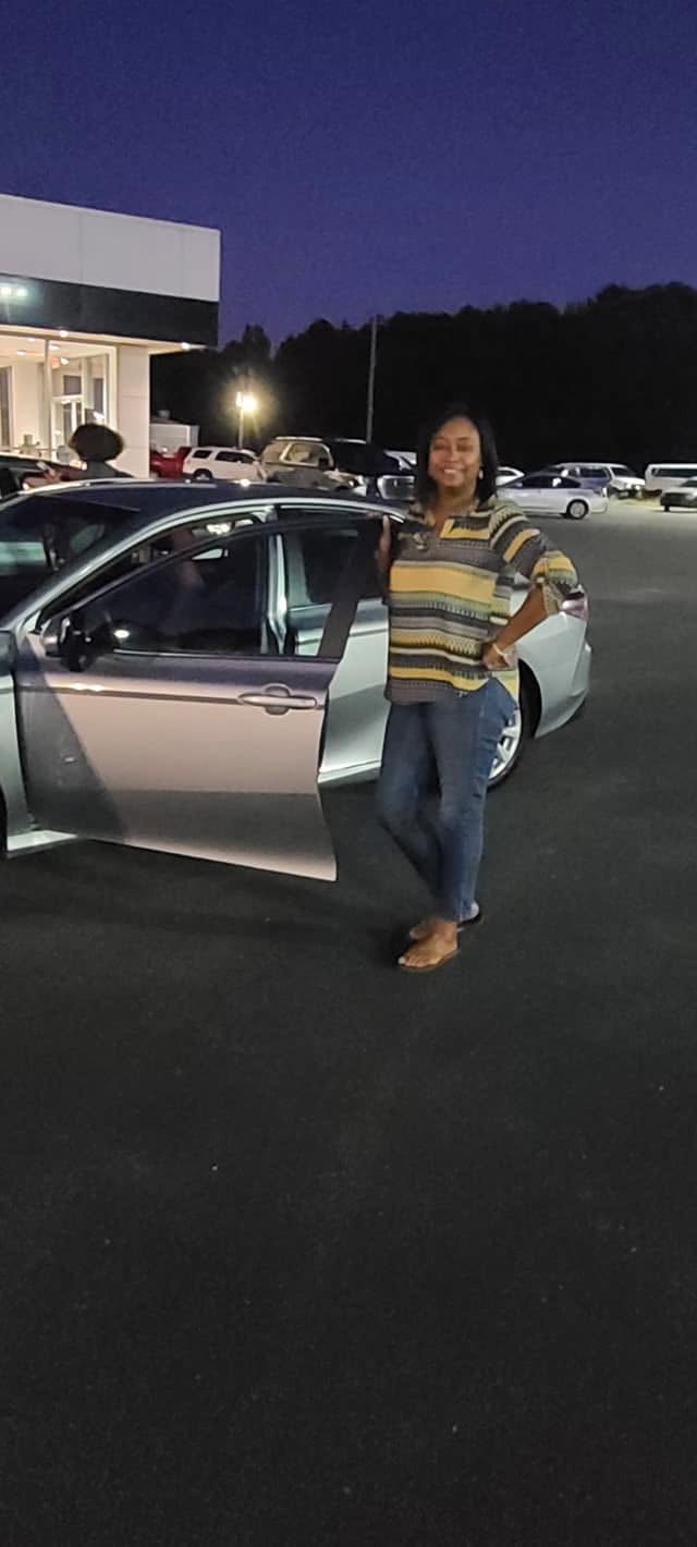 Woman standing next to a white sedan in a parking lot at dusk with a dealership building in the background
