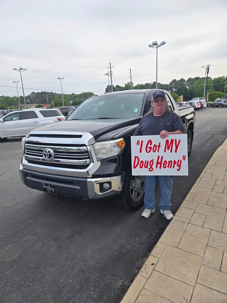 Man holding sign I Got MY Doug Henry stands next to black Toyota Tundra pickup truck in dealership parking lot