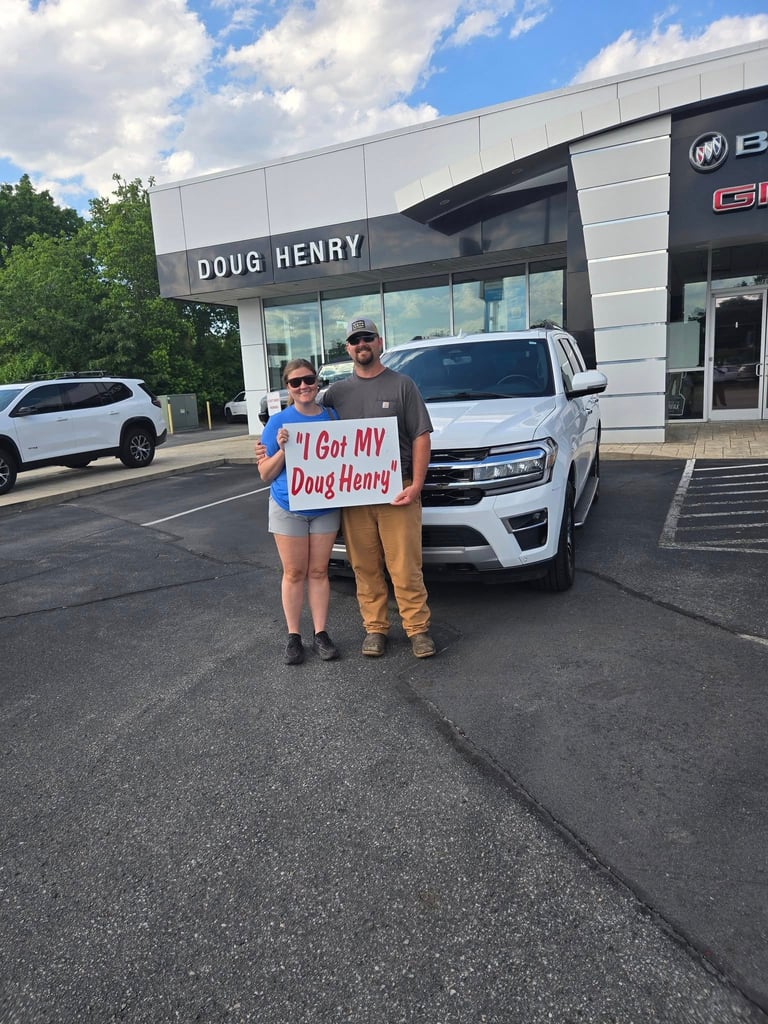 Two people holding a sign that says I Got My Doug Henry at a car dealership with white vehicles and storefront