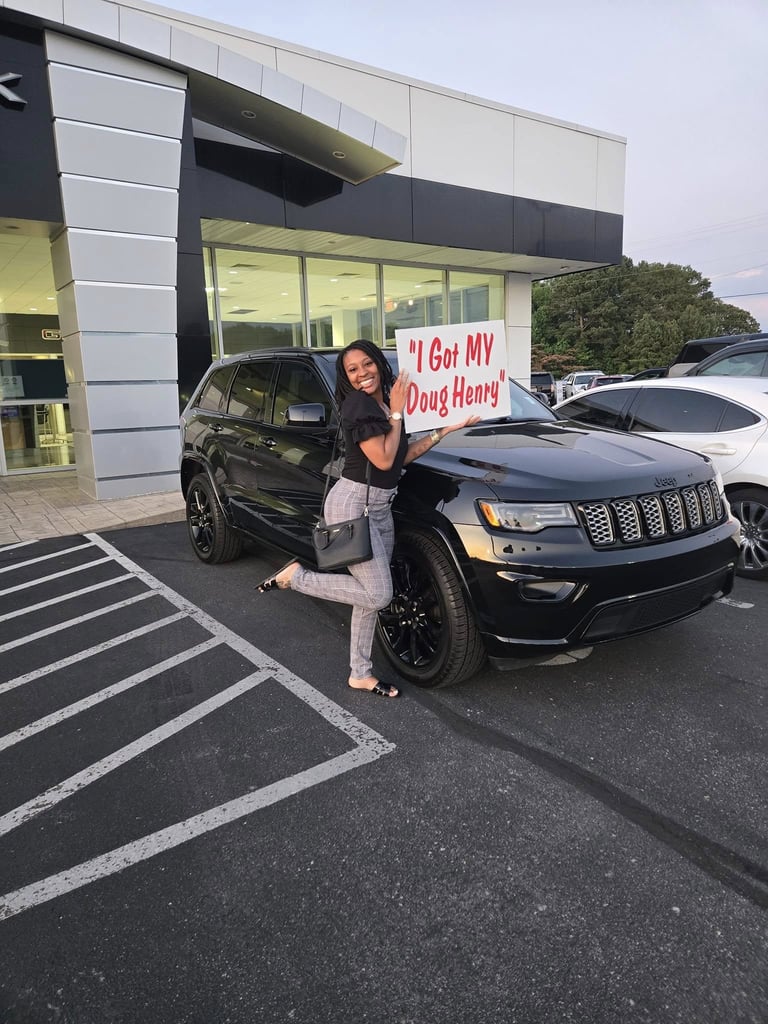 Woman posing next to a black Jeep Grand Cherokee in a dealership lot, holding a sign reading I Got MY Doug Henry