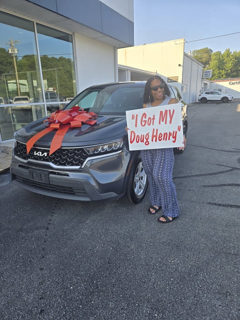Woman holding sign reading I Got MY Doug Henry standing next to new silver Kia SUV with red gift bow at dealership