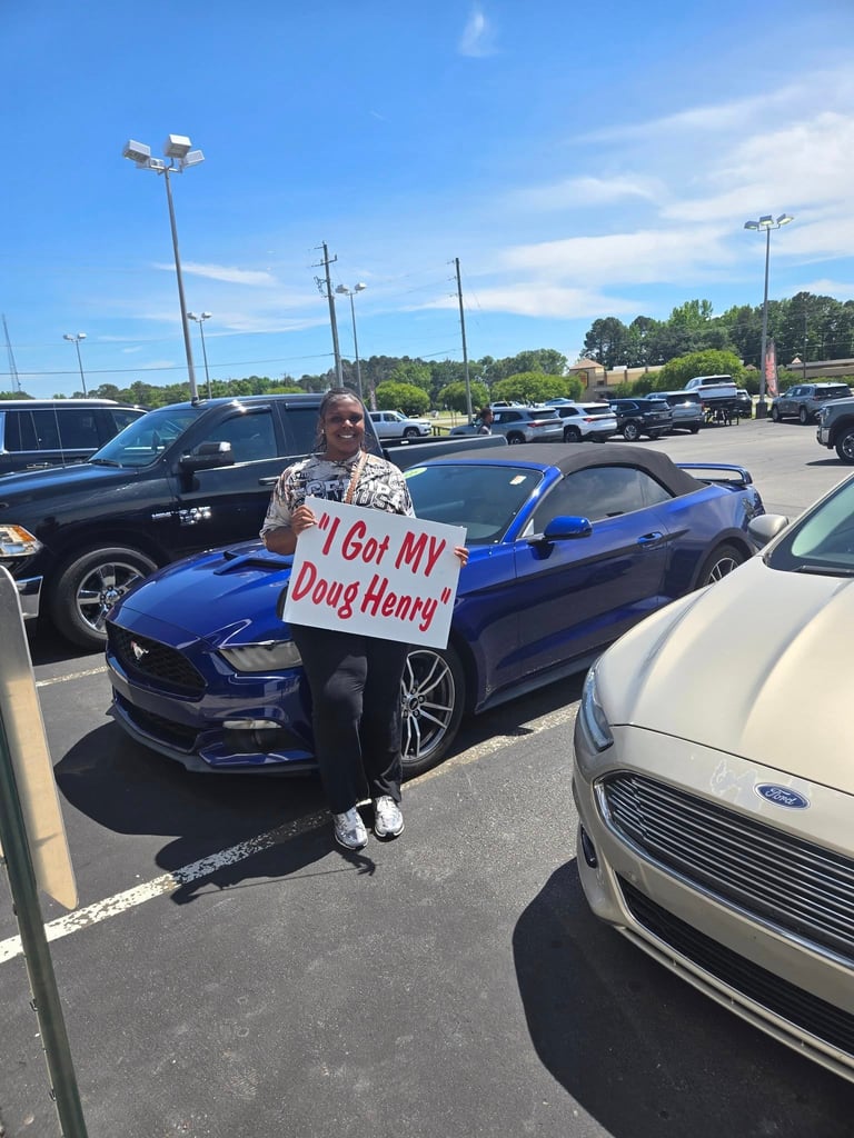 Man standing next to blue Mustang convertible holding sign reading I Got MY Doug Henry in parking lot