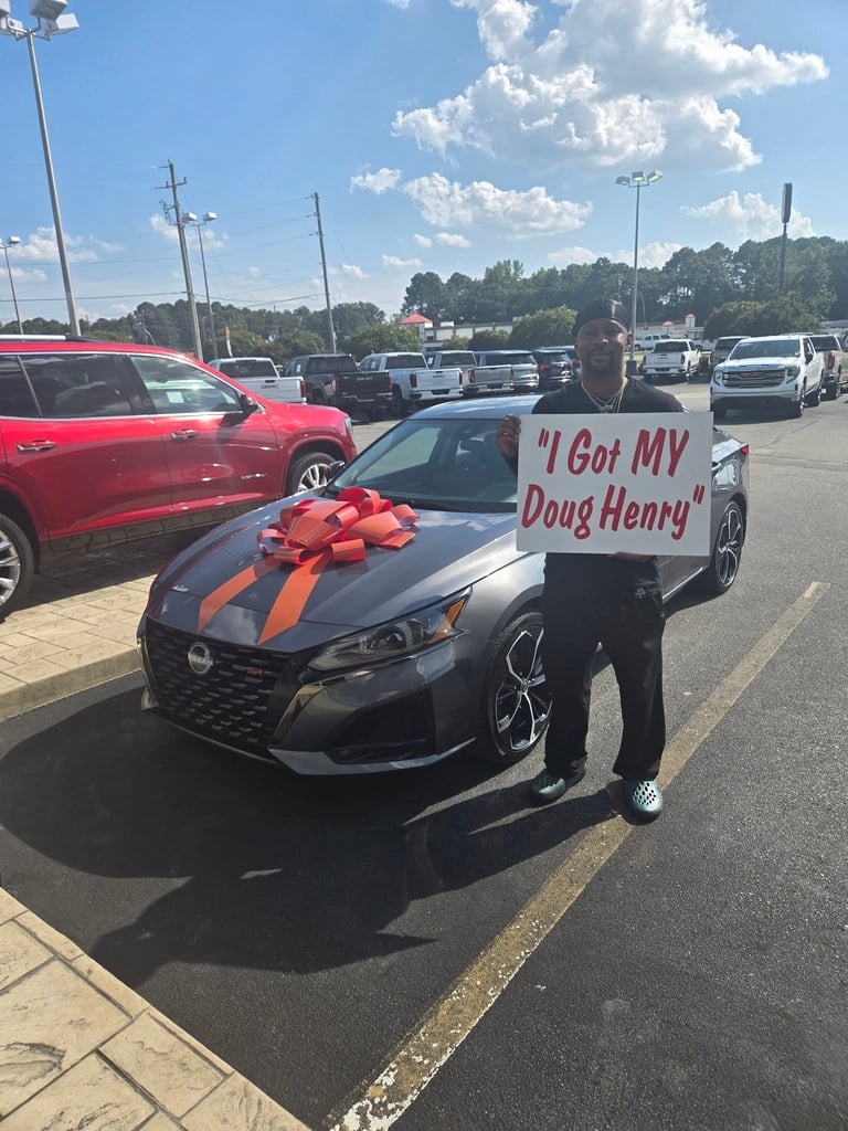 Person holding sign next to black car with large red bow in dealership lot, sign reads I Got My Doug Henry