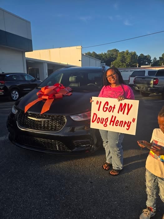 Woman holding sign I Got MY Doug Henry standing next to black car with red bow in dealership lot
