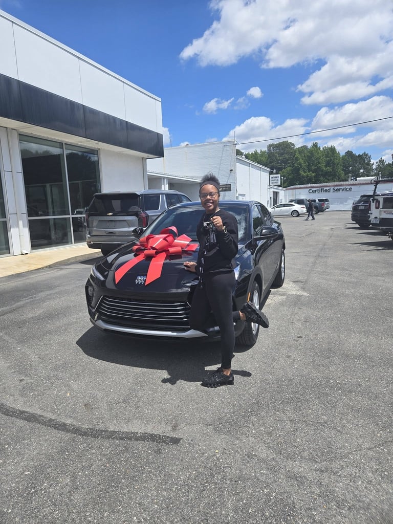 Person standing next to a black car with a large red bow at a car dealership lot