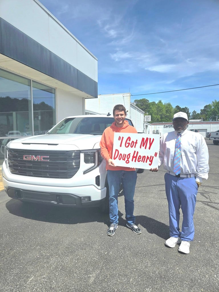 Two men stand in front of a white GMC pickup truck at a dealership, holding a sign that reads I Got MY Doug Henry