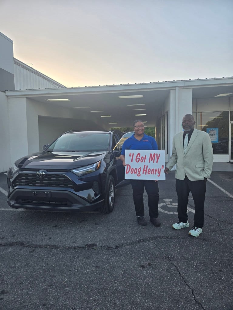 Two men standing beside a black Toyota RAV4 at a dealership, one holding a sign that reads I Got MY Doug Henry