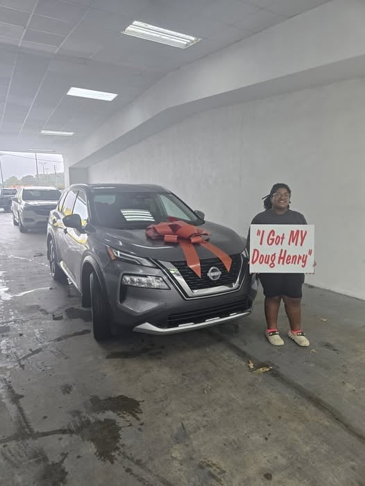Person holding sign reading I Got MY Doug Henry next to black Nissan with red bow in parking garage